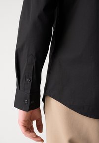 Camicia nera con maniche lunghe, caratterizzata da un polsino a punta, due bottoni e una texture liscia; indossata sopra un indumento beige.