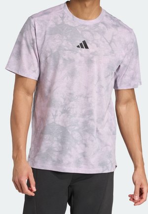 Mann trägt ein helllila Batik-Kurzarmshirt mit kleinem schwarzem Adidas-Logo auf der Brust und schwarze Hose.