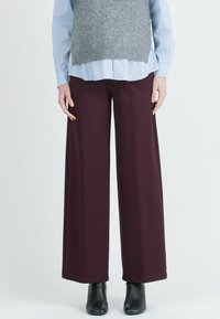 Wijduitlopende, bordeauxrode broeken van rekbaar materiaal, gecombineerd met een grijze mouwloze top over een blauw gestreept shirt. Zwarte enkellaarsjes zichtbaar.