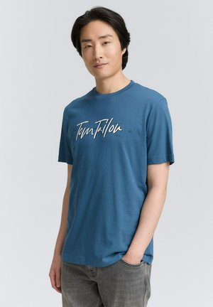 Camiseta azul de cuello redondo, hecha de algodón suave, presenta un gráfico de texto en blanco en cursiva "Tom Tailor", con un ajuste holgado y mangas cortas.