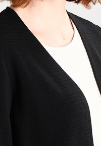 Černý texturovaný kardigan s otevřeným předním dílem, vyrobený z měkké, mačkané látky a s volným střihem. Nošený přes bílý žebrovaný top.