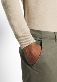 Gebreide trui in crèmekleur met ribbels aan de mouwen, gecombineerd met olijfgroene broek met zijzakken. Zichtbare hand in de zak detail.