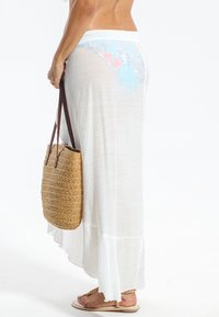 Jupe maxi blanche et fluide avec un design à niveaux, tissu texturé et aspect léger et transparent, associée à un sac fourre-tout en paille tressée.