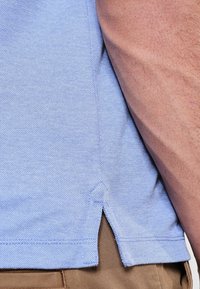 Polo bleu clair avec un tissage texturé, des fentes latérales à l'ourlet et une finition lisse. Le tissu semble respirant et léger.