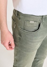 Pantaloni di cotone verdi con vestibilità dritta, dotati di tasche laterali e posteriori, e una piccola etichetta sulla tasca posteriore. Texture liscia.