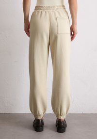 Beige Trainingshose mit elastischem Bund und Bündchen an den Knöcheln. Eine Gesäßtasche mit gesticktem Detail. Glatter Stoff, lockere Passform.