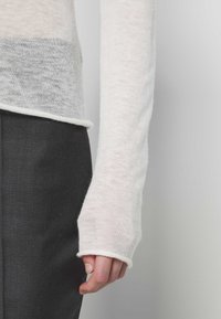 Gros plan sur la main et l'avant-bras d'une personne portant un haut léger, transparent à manches longues, associé à un pantalon foncé et ajusté sur un fond uni.