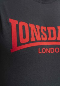 Sötétszürke póló, élénk piros "Lonsdale" felirattal és ívelt aláhúzással, sima anyagból készült, kerek nyakú dizájnnal.