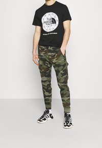 Musta puuvillainen T-paita, jossa valkoinen geometrinen logo, yhdistetty vihreisiin maastokuviollisiin cargo-housuihin, joissa on useita taskuja ja kapeahko leikkaus. Mustat lenkkitossut.