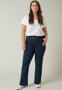 Jeans a zampa d'elefante in denim blu scuro con cinque tasche e vita alta, abbinati a una t-shirt bianca a maniche corte con scollatura a V e sneakers bianche.