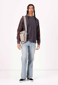 Grå langærmet skjorte, lyseblå denimjeans og sorte sneakers. Modellen holder en grå crossbody-taske med en lynlåslomme, mens hun står foran en hvid baggrund.