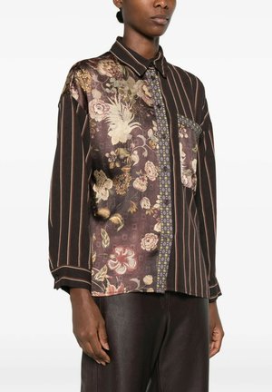 Donna che indossa una camicia a maniche lunghe con bottoni, con motivi floreali misti e righe verticali, abbinata a pantaloni in pelle scura.