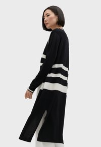 Lång svart kofta med vita horisontella ränder, tillverkad av stickat tyg, med långa ärmar och sidoslitsar för ökad rörelsefrihet.