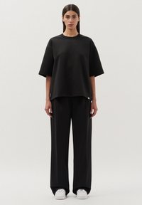 Zwarte oversized top met korte mouwen, gecombineerd met losse zwarte broek en witte sneakers. Glad materiaal, minimalistisch ontwerp, zonder patronen of accenten.