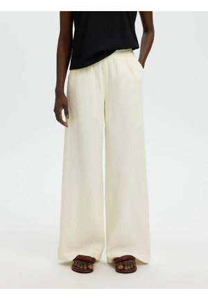 Personne portant un pantalon large et ample couleur crème, un haut noir sans manches et des sandales marron, debout avec une main dans la poche.