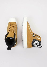 Zapatillas altas de ante marrón con puntera de goma blanca, acentos negros en el tobillo, ojetes plateados y un parche de logo en el lado exterior.