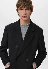 Joven con cabello castaño ondulado que lleva un abrigo negro de doble botonadura sobre un suéter de punto gris, con las manos en los bolsillos del abrigo, fondo neutro.