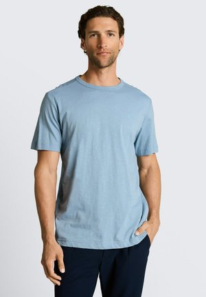 Mężczyzna o krótkich kręconych włosach, ubrany w jasnoniebieski t-shirt z krótkim rękawem i ciemne spodnie, stoi z jedną ręką w kieszeni na jednolitym tle.