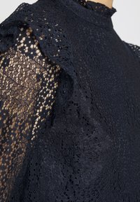 Námořnická krajková blůza s volánkovými rukávy, která má složité vzory a vysoký límec. Lehké provedení s průsvitným prvkem na rukávech.