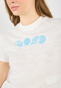 Balts T-krekls ar apaļu kakla izgriezumu, ar zilu čaulas grafiku uz krūtīm. Viegls, mīksts audums ar smalkām svītrām.