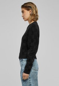 Zwarte gebreide trui met een gestructureerd geruit patroon, ribbels aan de mouwen en onderkant. Gepaard met lichtblauwe denim jeans.