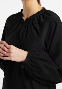 Blouse noire avec un col froncé et des manches bouffantes à poignets élastiques, fabriquée dans un tissu lisse et léger.