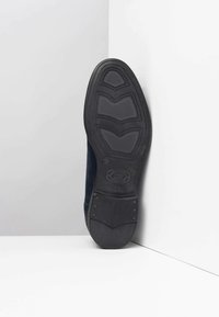 Suola di scarpa in gomma nera con grip texturizzato. Presenta scanalature curve e logo in rilievo. Fianchi realizzati in materiale scamosciato scuro.