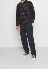 Camisa de cuadros en azul marino, verde y naranja, combinada con pantalones oscuros y zapatillas negras. Cuenta con mangas largas y un collar clásico.