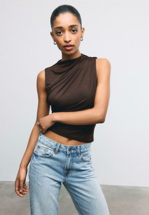 Femme aux cheveux foncés tirés en arrière, portant un haut marron sans manches et un jean bleu clair, debout avec un bras croisé sur sa taille.