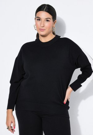 Kvinde med mørkt hår i lav hestehale iført sort sweater med hvid stribe på ærmet og sorte bukser, poserer med hånd på hoften.