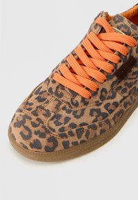 Tenisice sa uzorkom leopard, smešnog gornjeg dela od smeđeg antilopa, narančastim teksturiranim vezicama i gumiranim đonom. Detalji uključuju kontrastno šivenje i zaobljen nos.