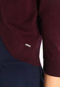 Närbild av en person som bär en ribbad vinröd tröja med en liten etikett "More & More" nära fållen och marinblå byxor.