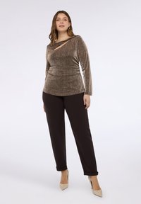 Braunes, strukturiertes Langarmshirt mit einem diagonalen Ausschnitt, kombiniert mit dunkelbraunen Hosen und beigen Schuhen mit Absatz, vor hellem Hintergrund.