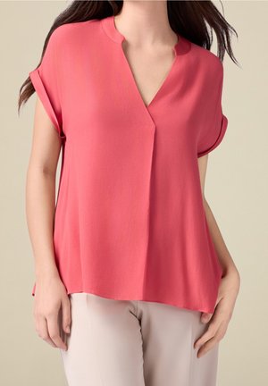 Frau trägt eine korallenfarbene Bluse mit kurzen Ärmeln, hochgekrempelten Manschetten und V-Ausschnitt, kombiniert mit hellbeigen Hosen vor neutralem Hintergrund.