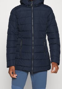 Doudoune marine avec un design matelassé horizontal, fermeture éclair intégrale à l'avant, deux poches latérales et une capuche amovible. Texture douce et lisse.