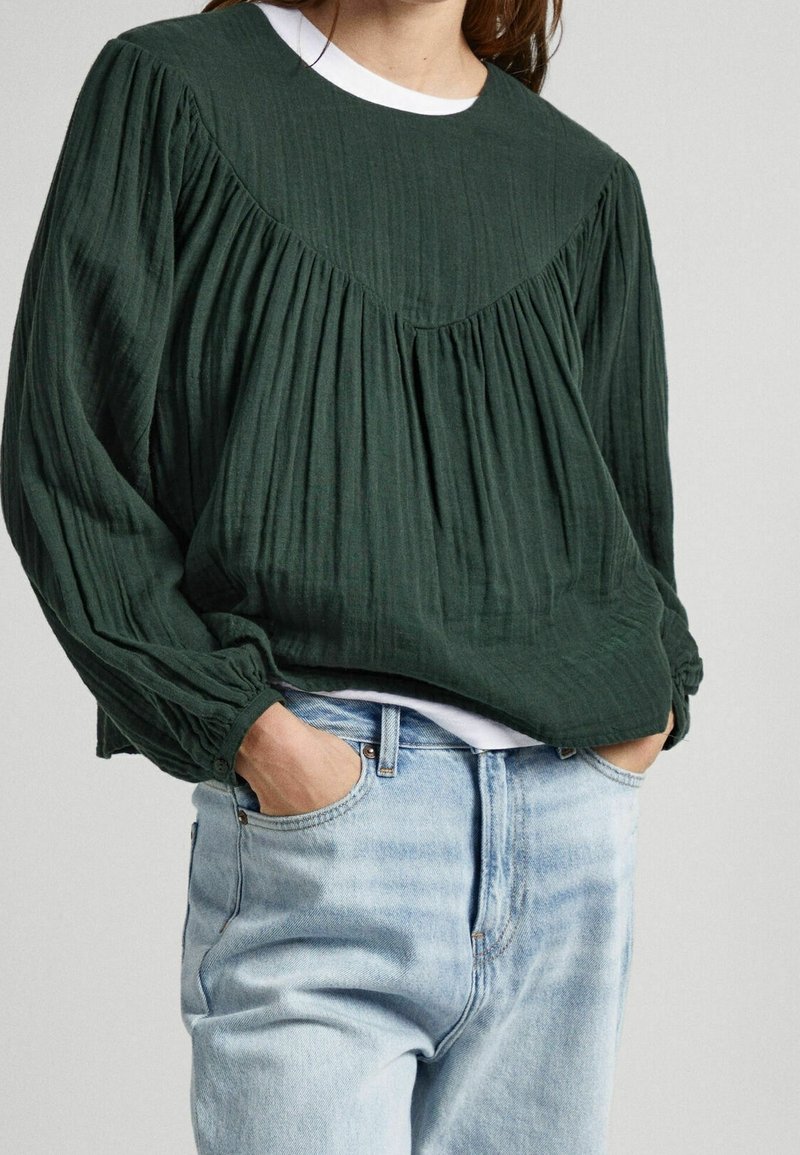 Femme portant un chemisier plissé à manches longues vert foncé sur un haut blanc, associé à un jean en denim bleu clair, les mains dans les poches.