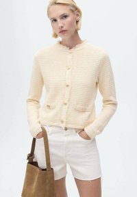 Cárdigan de textura color crema con un cuello deshilachado, cierre de botones y bolsillos frontales; combinado con pantalones cortos blancos y un bolso marrón.