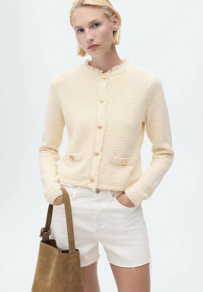 Cárdigan de textura color crema con un cuello deshilachado, cierre de botones y bolsillos frontales; combinado con pantalones cortos blancos y un bolso marrón.