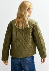 Chaqueta acolchada verde oliva con un patrón redondeado, que presenta mangas largas y un corte relajado, combinada con jeans de mezclilla azul.