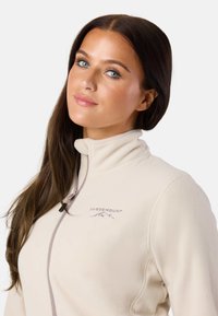 Beige fleece-takk med høy krage, med glidelås og brodert logo. Glatt tekstur og minimale designelementer.
