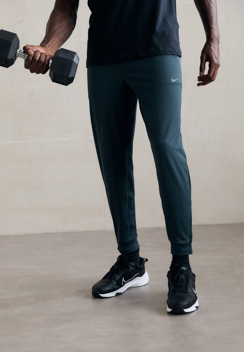 Spodnie sportowe Nike w ciemnozielonym kolorze, dopasowany krój z zwężającymi się nogawkami. Noszone z czarnymi butami sportowymi z białymi akcentami, trzymając hantel.