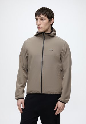 Ung mand iført en beige hættetrøje med lynlås og BOSS-logo, står mod en ensfarvet hvid baggrund og kigger til siden.