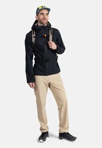 Zwarte waterdichte jas met verstelbare capuchon, beige broek en zwarte sneakers. Inclusief een rugzak met bruine banden en een oranje clip.