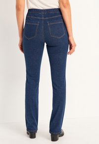 Kvinde iført mørkeblå denimjeans med elastisk talje og lige ben, stående med ryggen mod kameraet på en ensartet hvid baggrund.