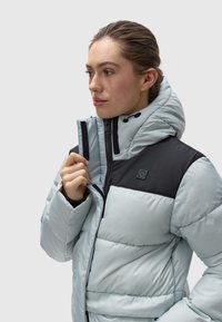 Pūkuota striukė šviesiai mėlynos spalvos su juodais akcentais. Yra aukšta apykaklė, užtrauktukas ir š quilting tekstūra. Logotipo etikete ant krūtinės.