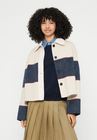 Κολάρο, cropped σακάκι σε ρίγες κρεμ, ναυτικό μπλε και βατόμουρου, με απαλή υφή και μαύρα κουμπιά. Φορεμένο πάνω από ένα ναυτικό μπλε πουλόβερ και φούστα.