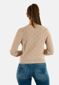 Suéter tejido beige con patrón de diamante, cuello redondo y mangas largas, combinado con jeans azules que presentan bolsillos traseros curvados.