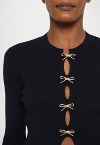 Gros plan d'une personne portant un pull noir côtelé avec quatre fermoirs dorés en forme de nœud sur l'ouverture avant.