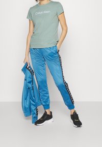 Camiseta de algodón verde claro con el logo de "Calvin Klein", combinada con pantalones de chándal satinados azul brillante con franjas laterales negras y zapatillas negras.