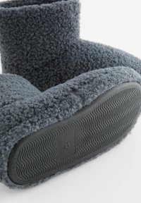 Graue Fleece-Hausschuhe mit einer weichen, strukturierten Oberfläche und einer dicken Gummisohle, die ein Wellenmuster für besseren Halt aufweist.